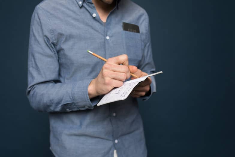 紺色の背景の前に立つ男性が鉛筆でノートに書いている