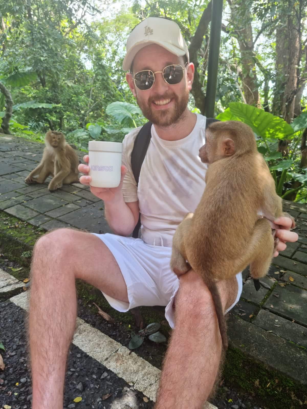 Un hombre sonriente con una gorra de béisbol blanca sentado en un ambiente tropical, sosteniendo una taza de bebida en su mano derecha y tocando un mono marrón que se balancea sobre su rodilla izquierda con su mano izquierda.