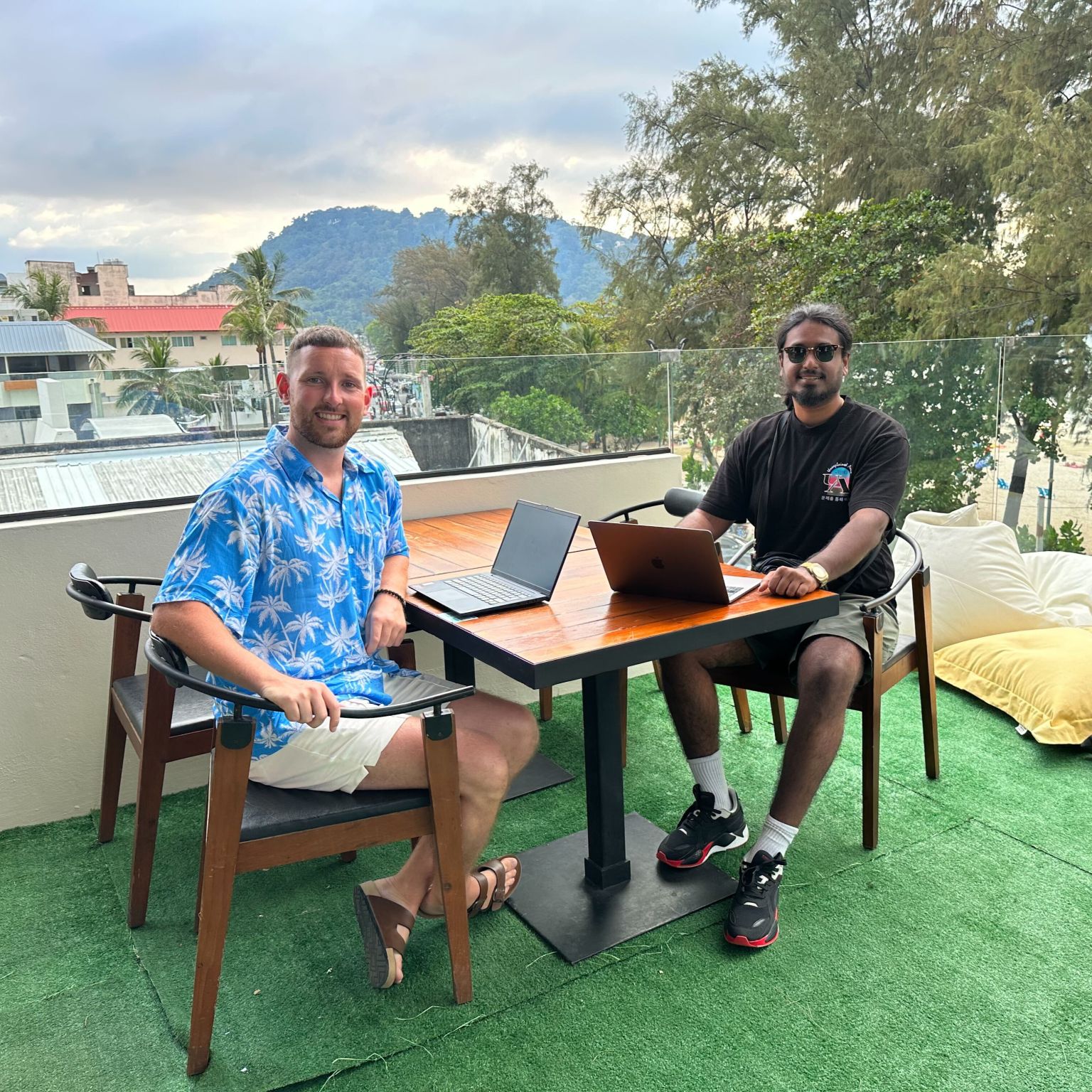 Dos hombres sonrientes sentados con computadoras portátiles en una mesa en una azotea al aire libre.