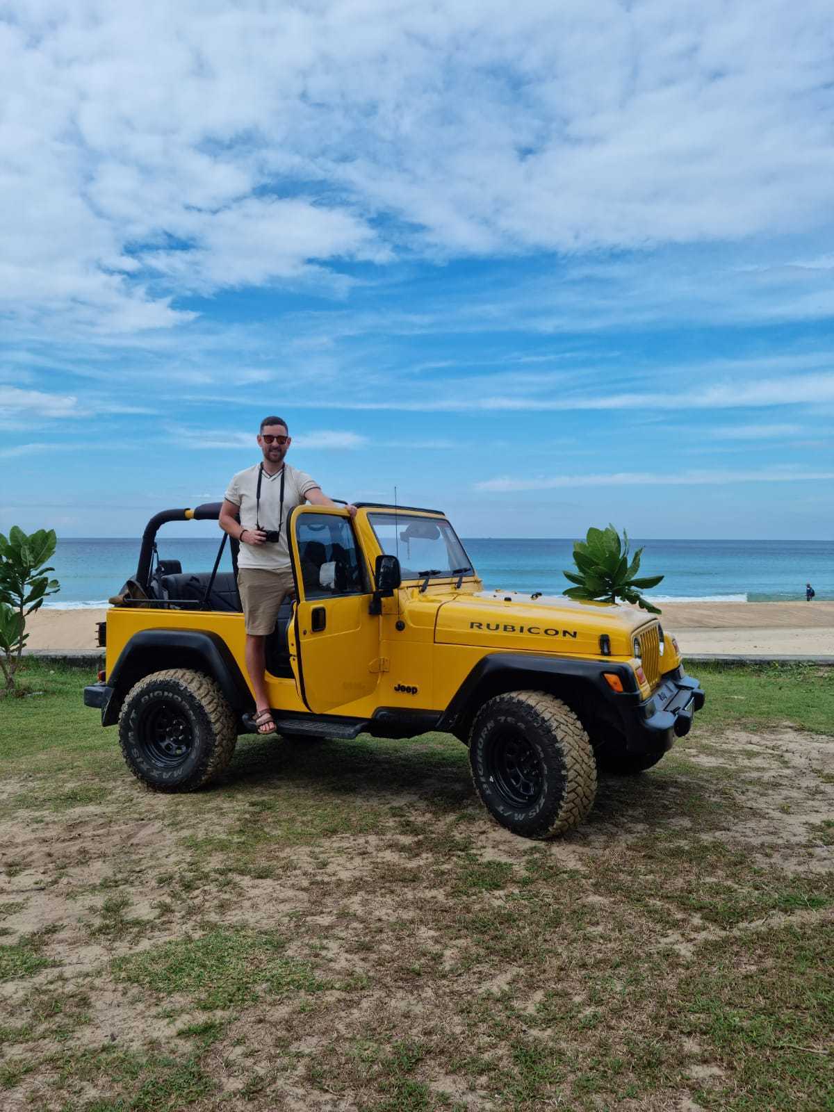 Un hombre parado en la puerta abierta de un Jeep Rubicon amarillo con vista al océano al fondo.