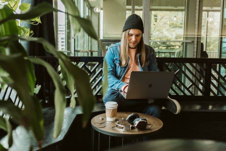 Mężczyzna pracuje na laptopie, siedząc przed małym stolikiem z kawą i słuchawkami