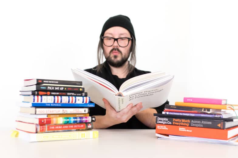 um homem de gorro lê um livro sobre design. duas pilhas de livros sentam-se em ambos os lados de sua mesa