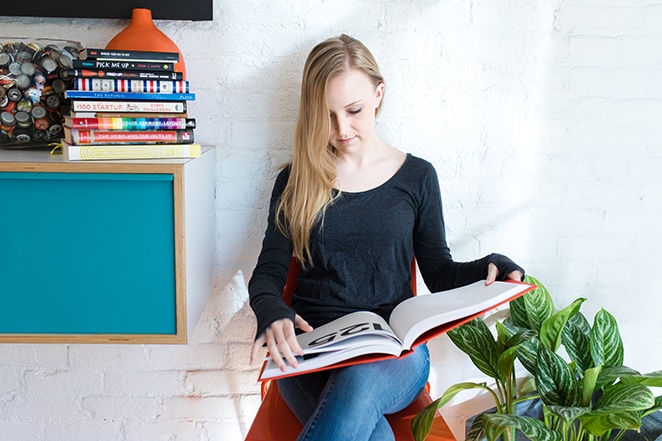 mujer en silla leyendo un libro de diseño con una pila de libros y una planta cercana