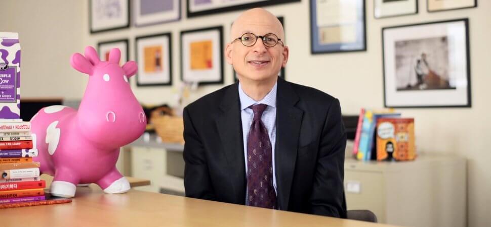 Image de Seth Godin où il est assis sur sa table de lecture avec une licorne et plein de livres autour de lui