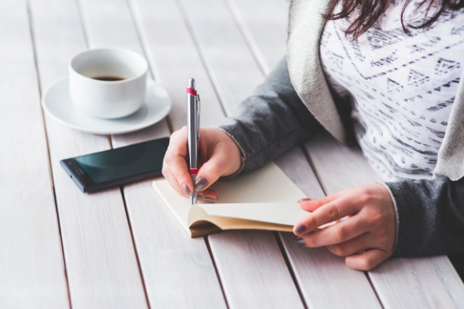 una mujer escribe en un cuaderno en un escritorio blanco al lado de su teléfono y una taza de café medio llena