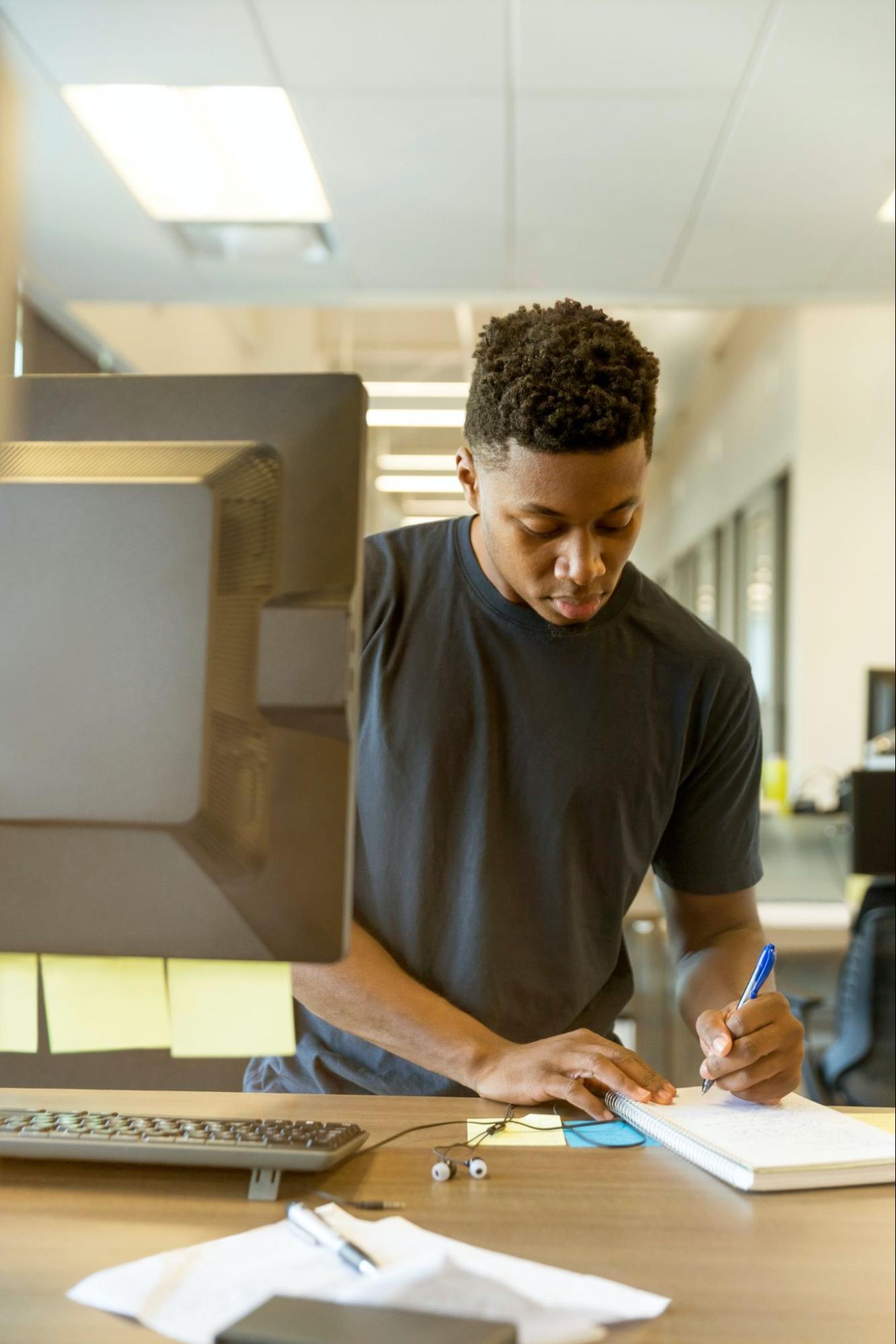 homem escrevendo em um bloco de notas ao lado de um computador