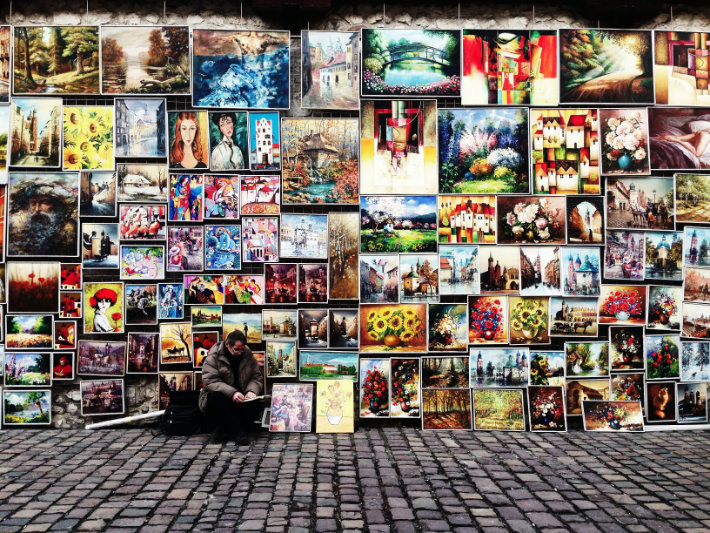 um homem com um casaco fofo está sentado em uma rua de tijolos em frente a uma parede totalmente coberta com diferentes obras de arte coloridas