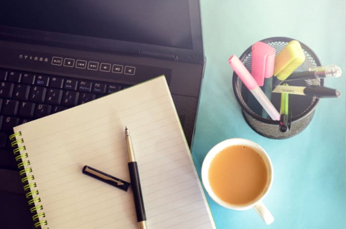 um laptop, caderno, caneta, porta-canetas cheio de utensílios de escrita e uma xícara de café em uma mesa azul