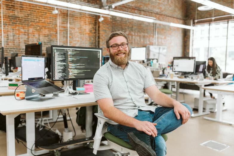 um homem barbudo sorri para a câmera enquanto está sentado em frente a um monitor de computador que exibe linhas de código