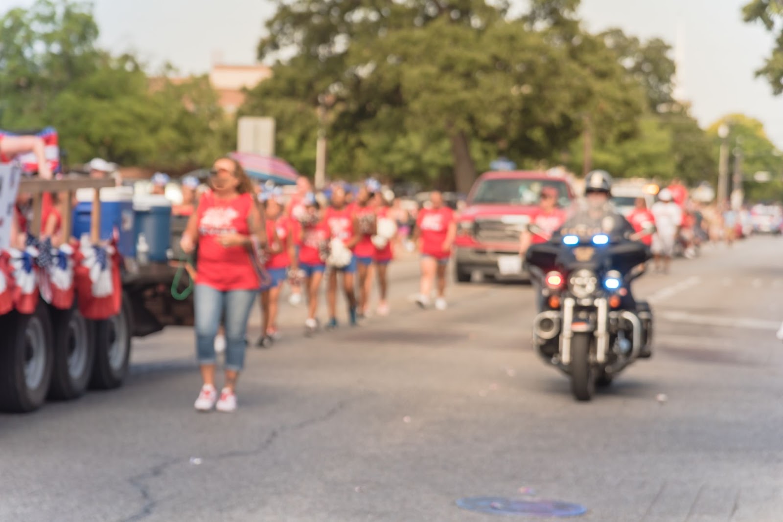 Verschwommenes Bild einer Parade am 4. Juli, bei der alle rote Hemden und blaue Hosen tragen, während ein Polizist auf einem Motorrad nebenher fährt.