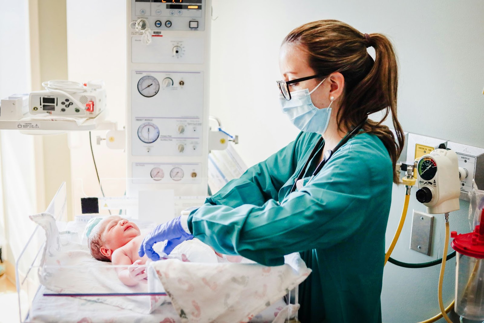 Bir bebeğin hayati değerlerini kontrol etmek için önlük, eldiven ve maske takan bir kadın doktor veya hemşire.