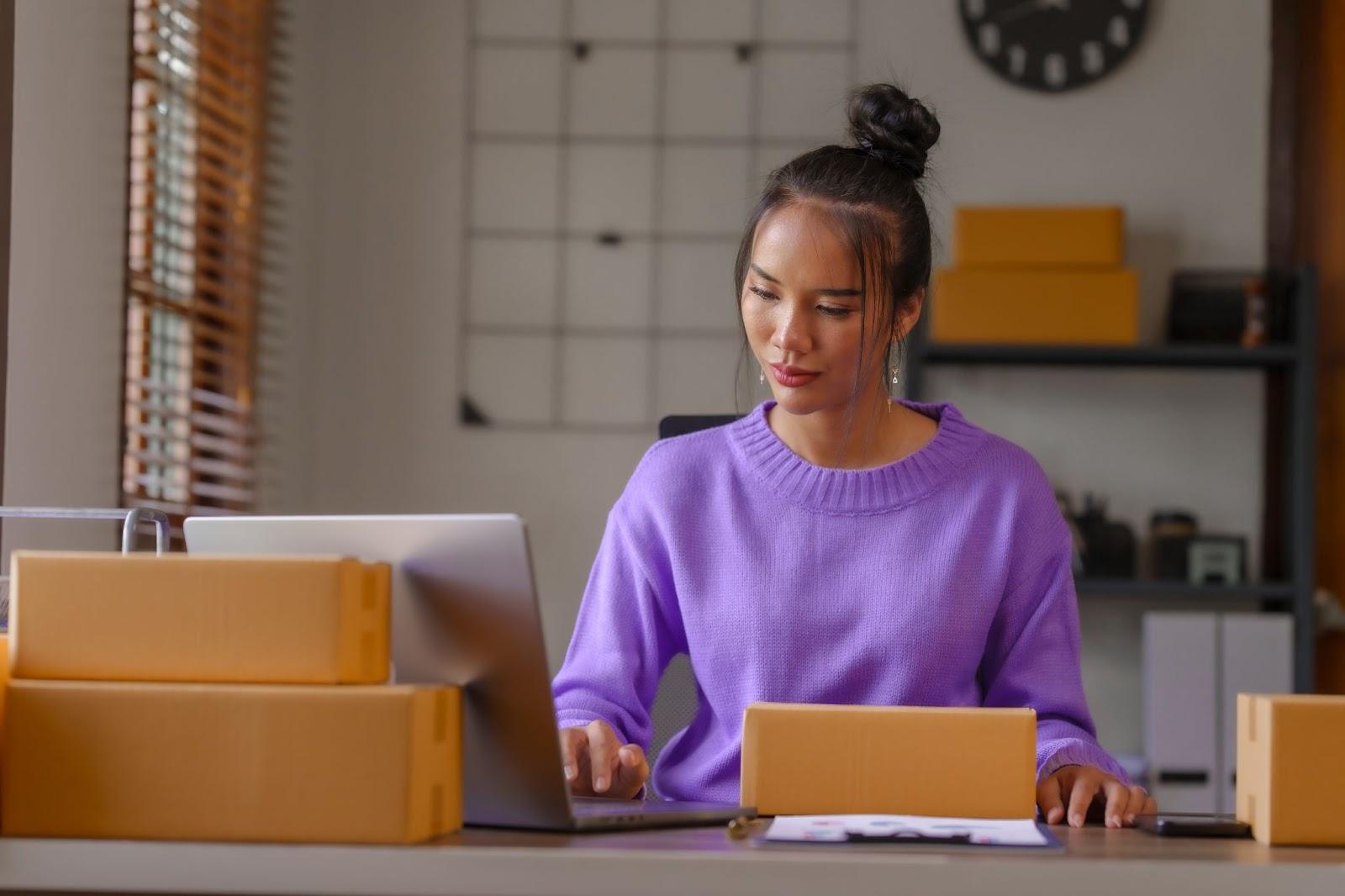 Mujer trabajando en su computadora en un escritorio en una oficina que parece estar preparada para la preparación de envíos.