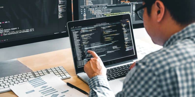フルスタック開発者はラップトップで作業し、フロントエンドとバックエンドのツールを表示する複数の画面でコーディングを行っています。